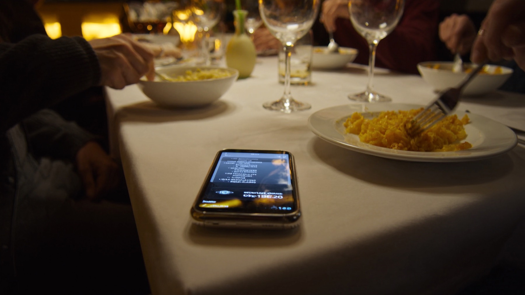 A phone face up on a restaurant table, screen lit, dinner in progress around it